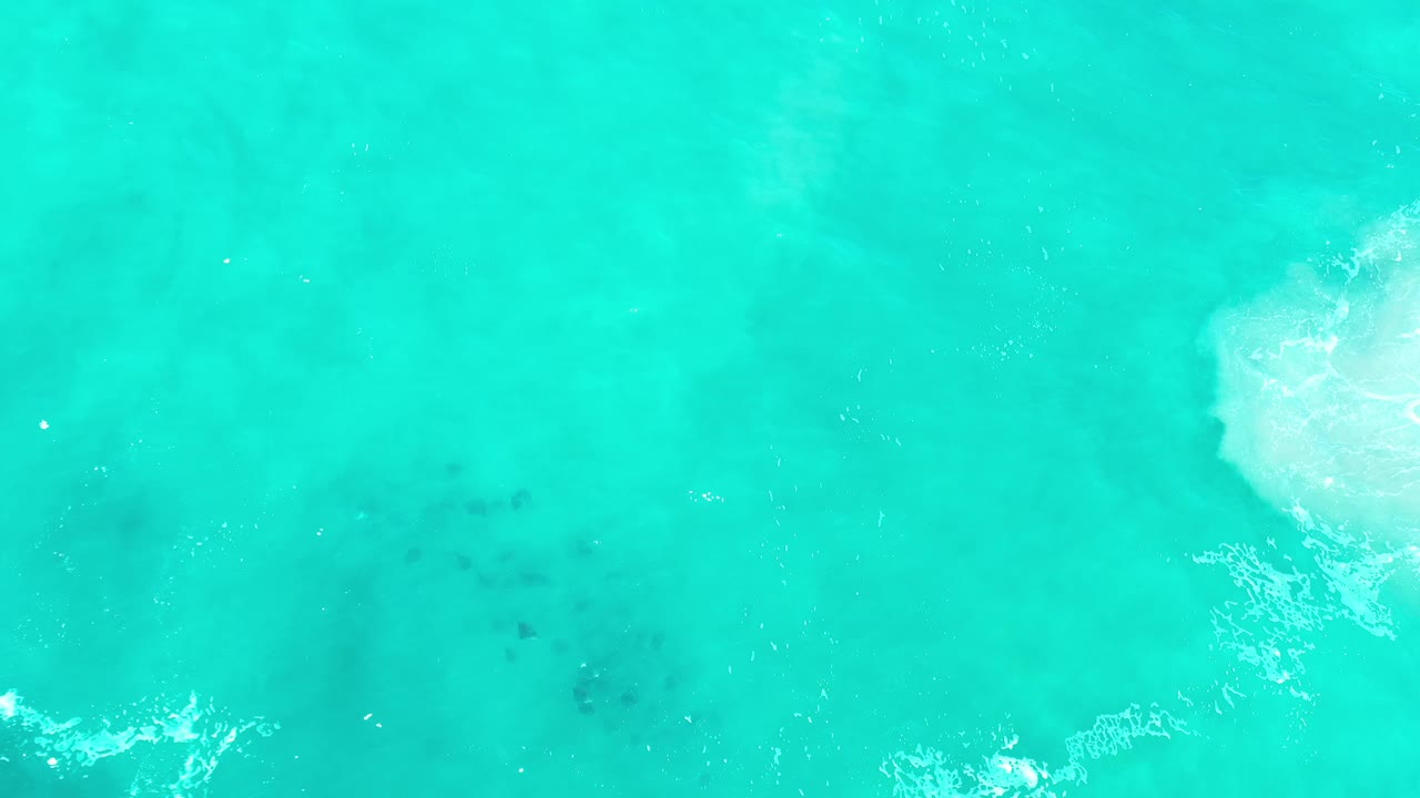 Aerial top-down ocean view in Los Cabos, Mexico, with a bright foamy patch near the upper right, muted darker water below, and no visible people.