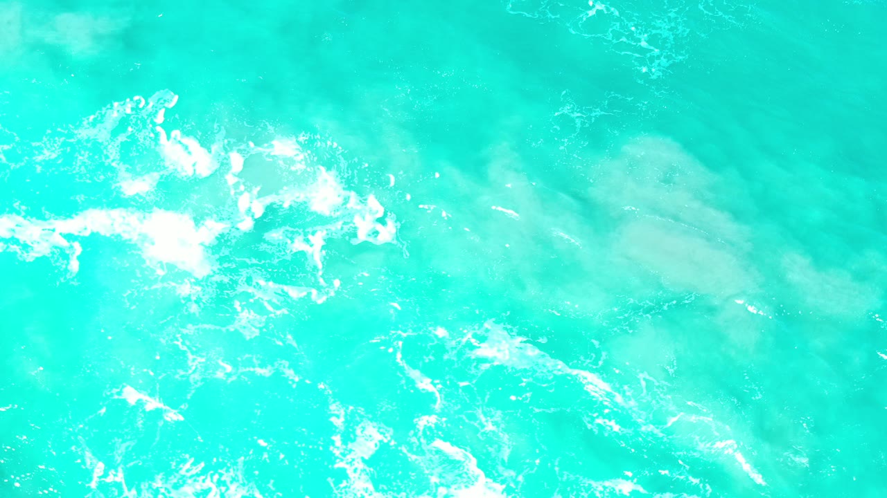 Overhead view of turquoise sea in Los Cabos, Mexico, with white foam near the left side, pale underwater shapes, and no visible people.