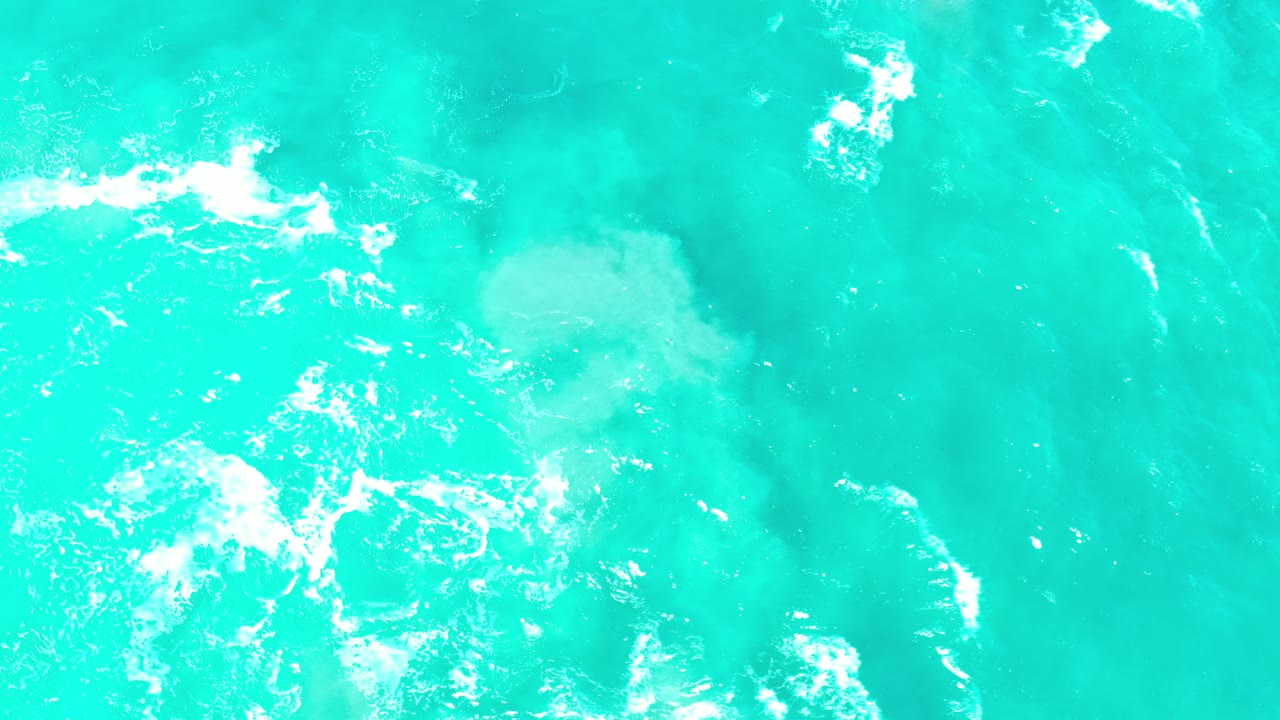 Top-down view of turquoise ocean water in Los Cabos with white foam patches near the left side and no visible people.