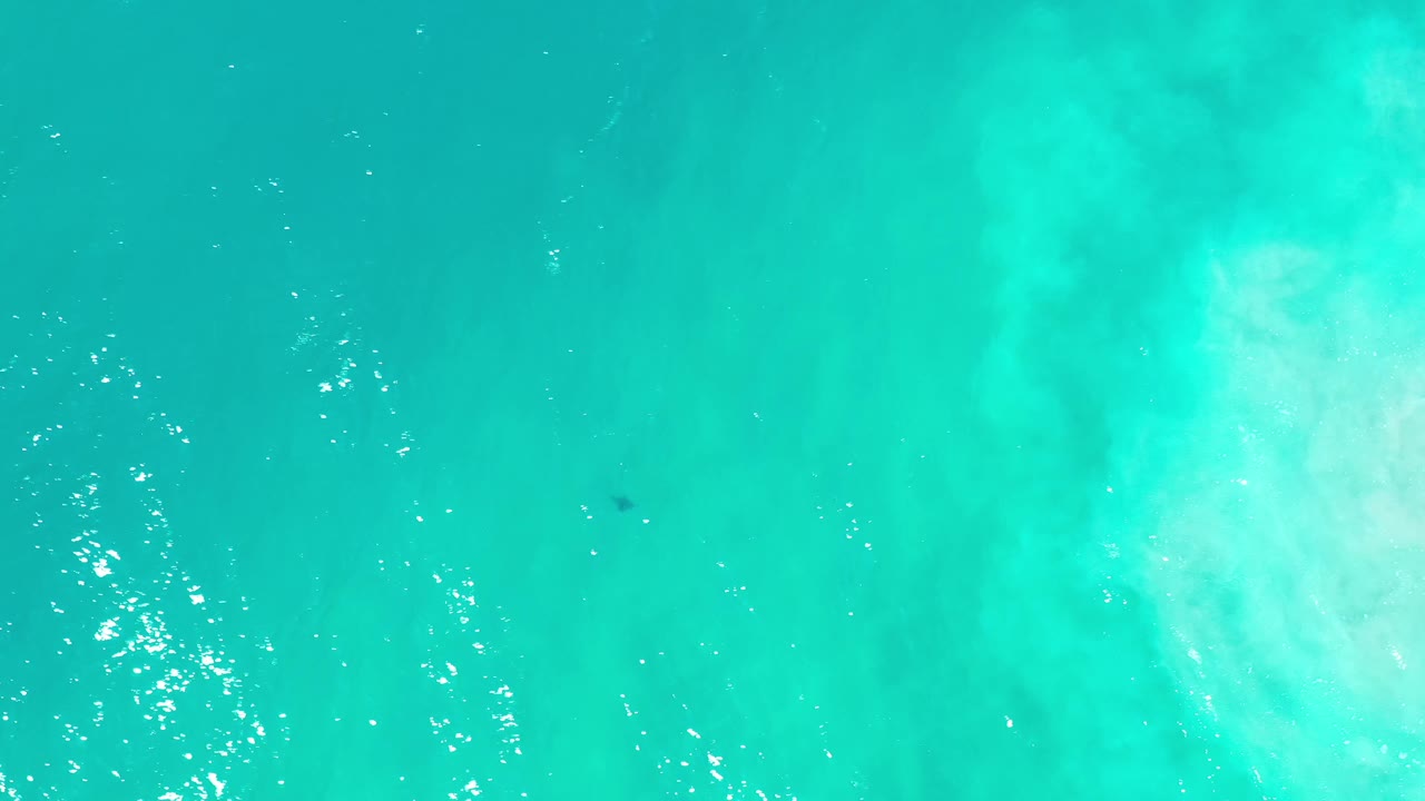 Aerial frame of clear blue-green Los Cabos ocean with bright surface ripples, a pale underwater patch near the lower left, and no visible people.