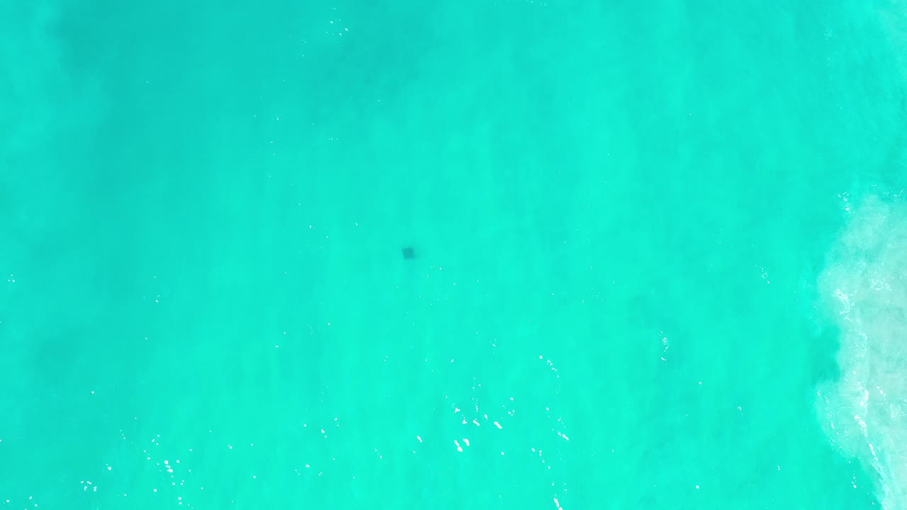 Overhead view of turquoise water in Los Cabos, Mexico, with a vertical strip of white surf near the right side and no visible people.