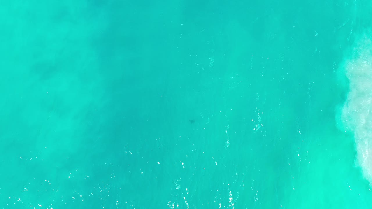 Top-down view of turquoise and green ocean water in Los Cabos, Mexico, with a white surf patch near the right edge and no visible people.