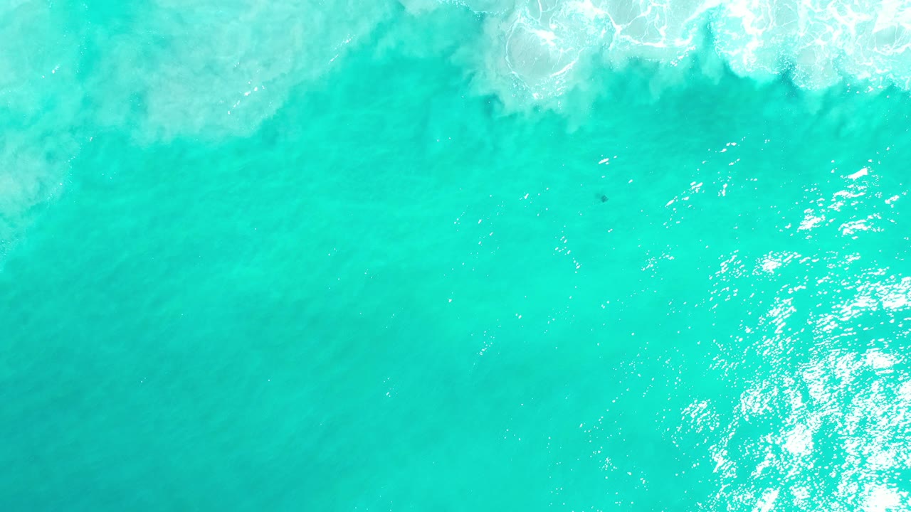 Overhead view of turquoise sea in Los Cabos with a band of white surf along the top edge and no visible people.