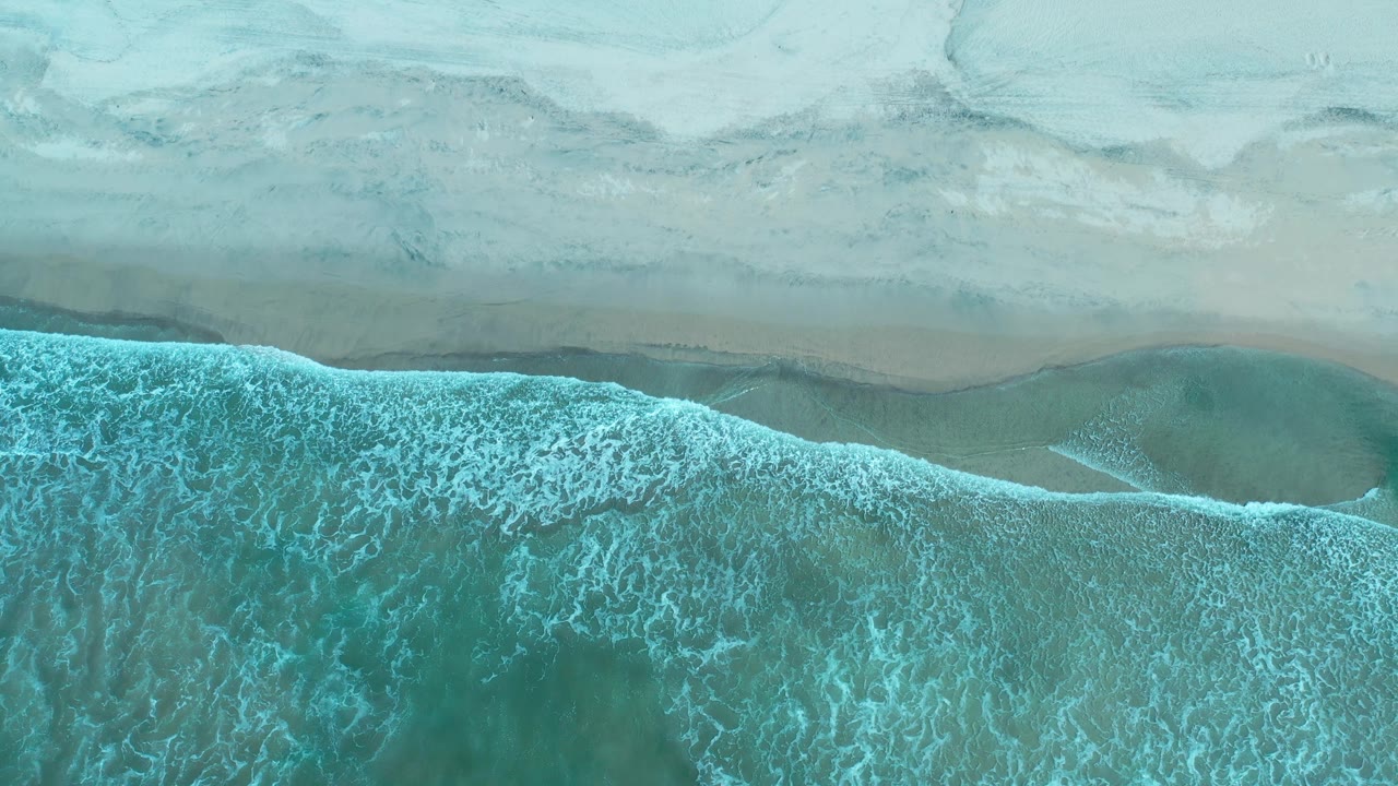 Top-down Los Cabos shoreline with a wide band of blue-green surf, wet sand, and no visible people.