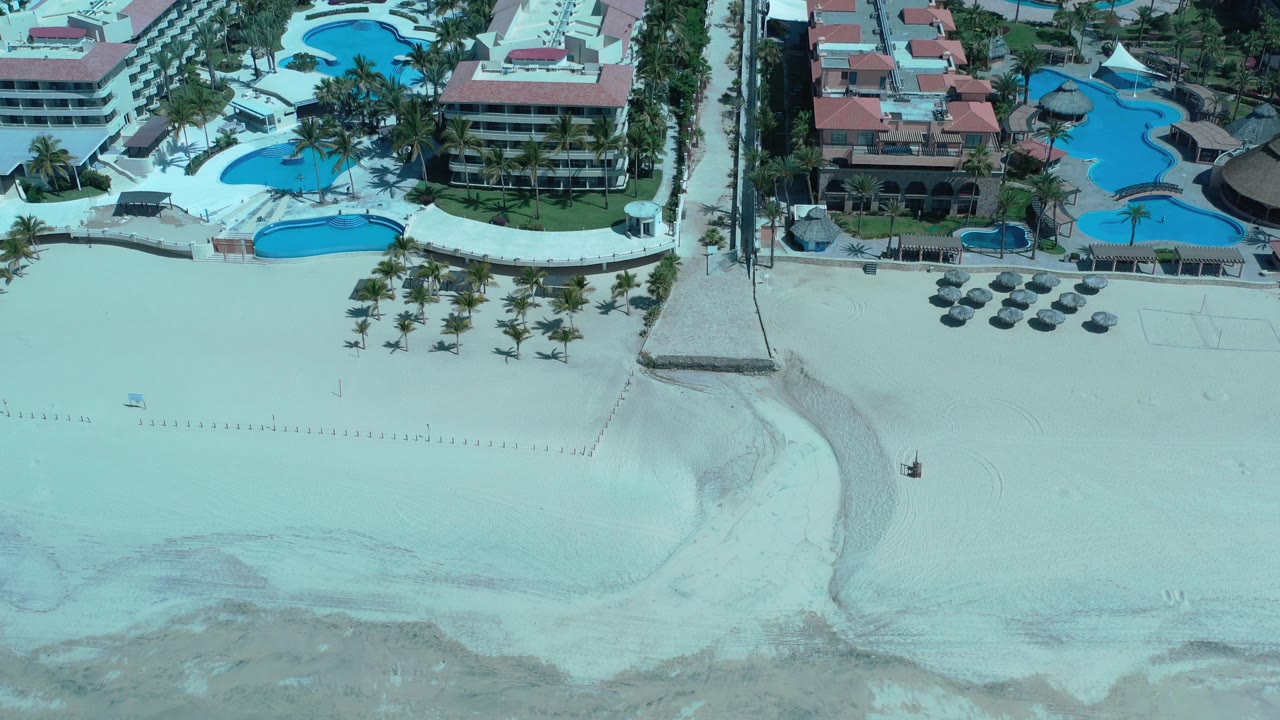 Aerial view of beachfront hotels, pools, palms, and pale sand.
