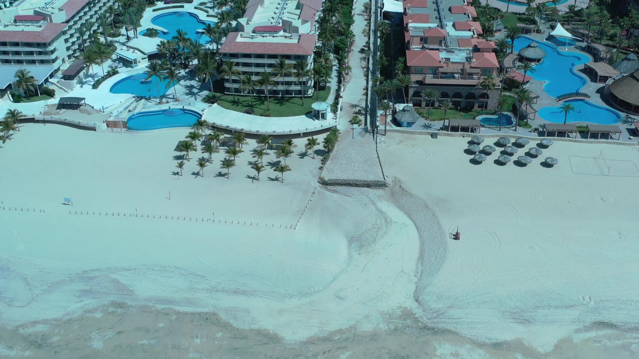 Aerial view in Los Cabos with the resort pools slightly more centered, showing a broad empty sand area, shoreline shallows, palms, umbrellas, and no visible people.