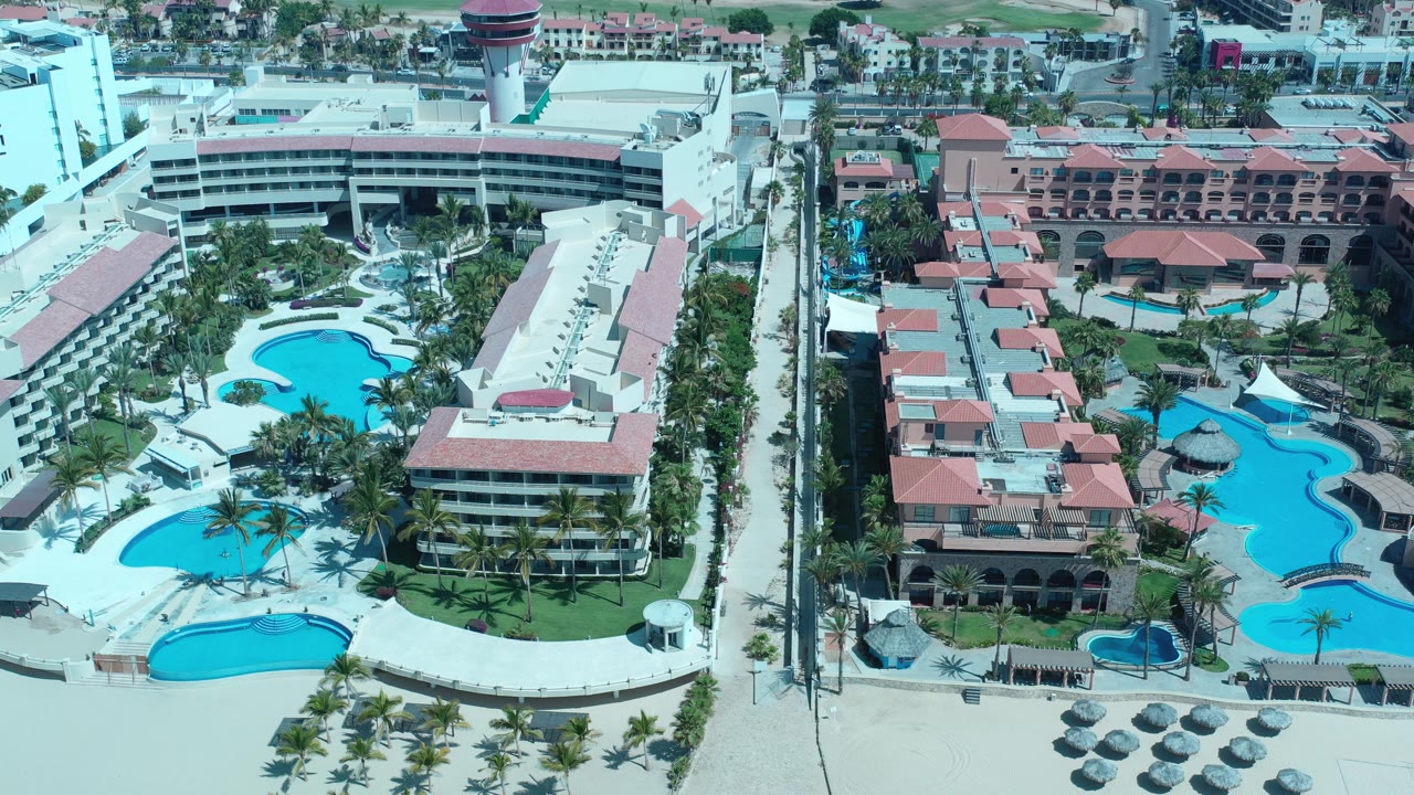 Aerial view over a Los Cabos resort with blue pools, palm landscaping, pale beach sand, red-roofed buildings, and tiny distant people near the grounds.