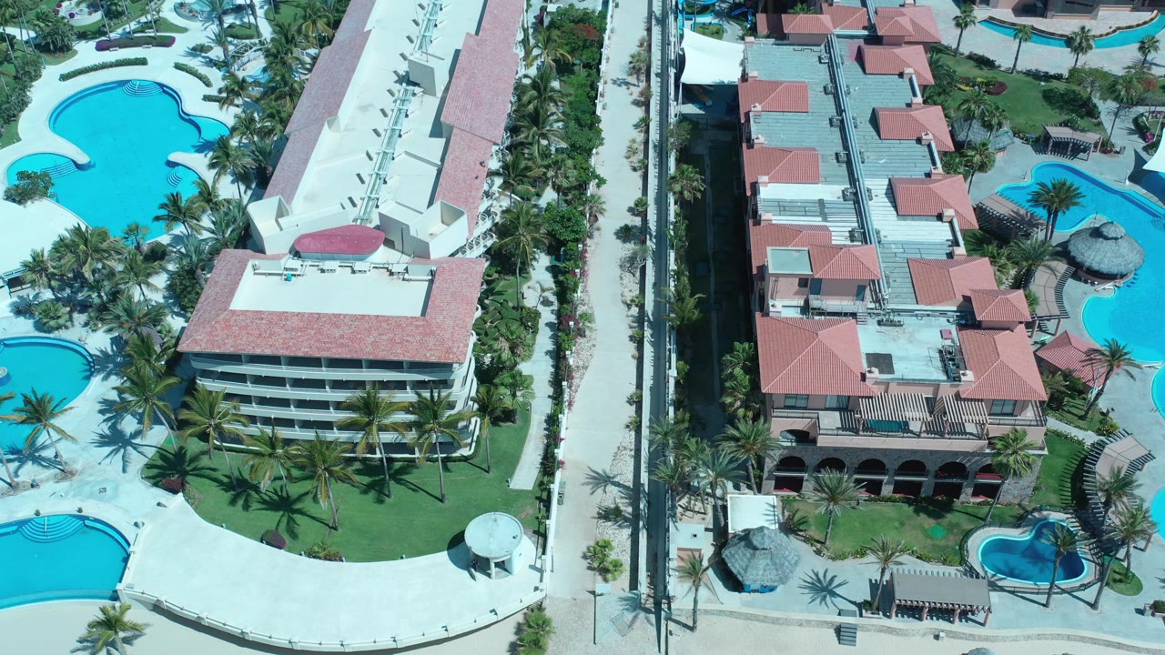 High aerial frame in Los Cabos, Mexico, with a white building on the left, red-roofed structures on the right, bright blue pools, palms, and no visible people.