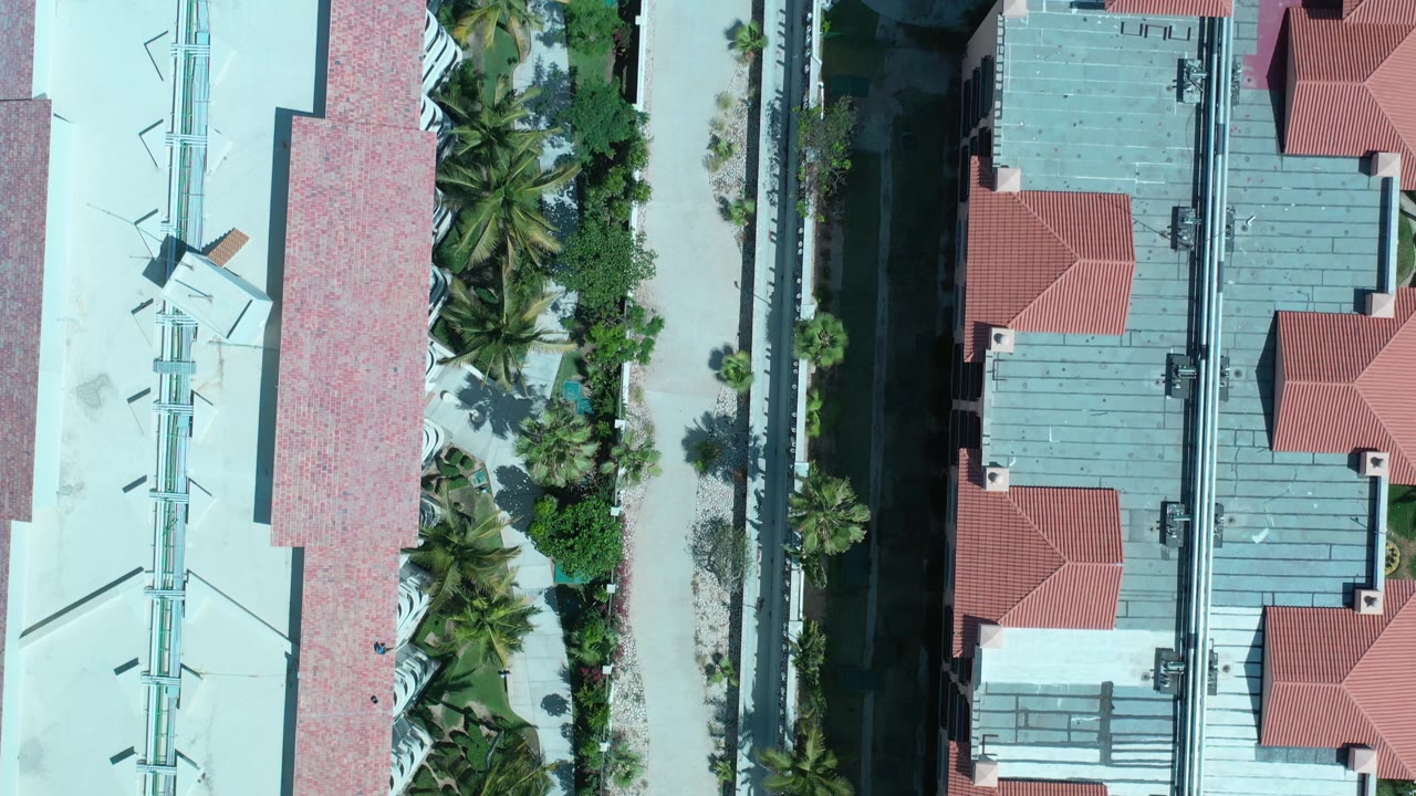 Overhead Los Cabos frame with red roof sections, gray roof panels, palm trees, planted strips, and a paved path along the buildings, with no visible people.