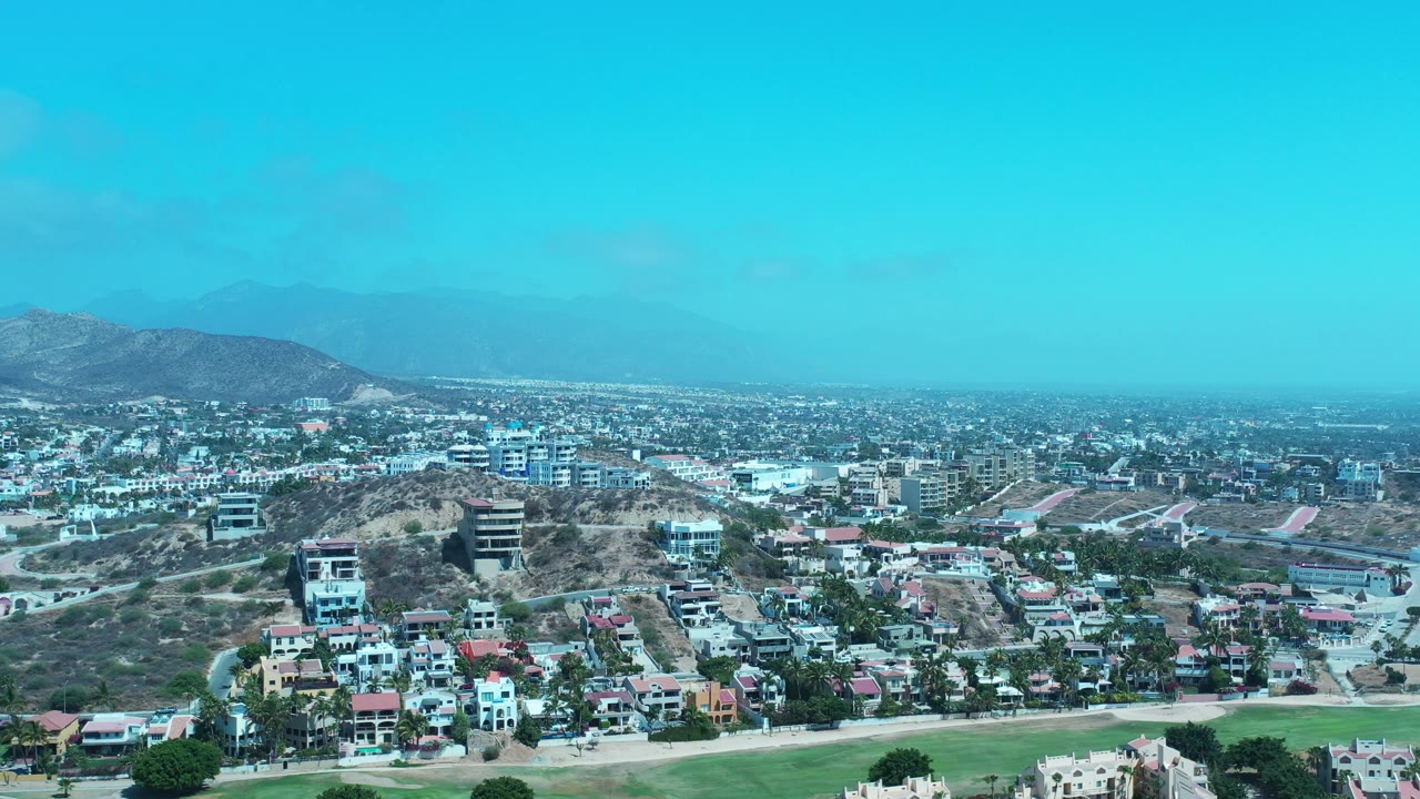 Wide aerial view over Los Cabos, Mexico, with hillside residences, low-rise city blocks, green foreground land, hazy mountains, bright blue sky, and no visible people.