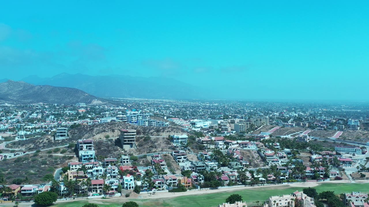 Los Cabos aerial panorama showing a rounded hillside with homes and buildings in front of a wide urban area, distant mountains, blue sky, and no visible people.