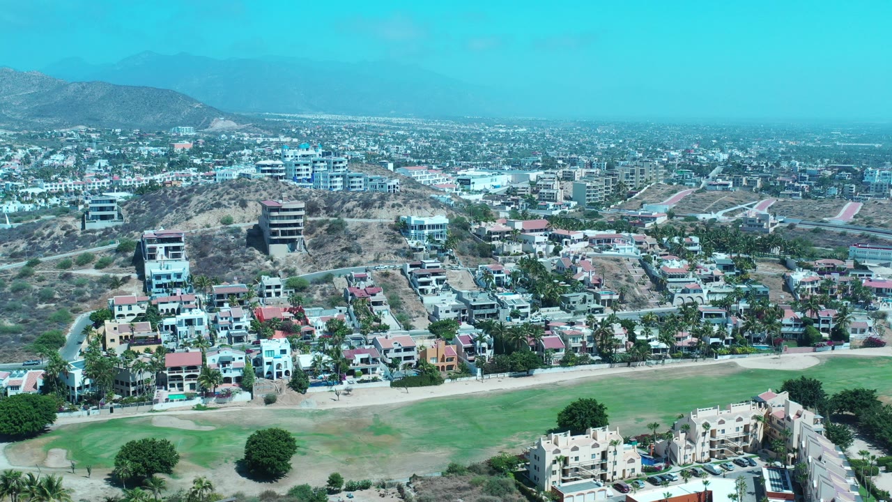 Aerial view of a golf course below hillside homes and city buildings.