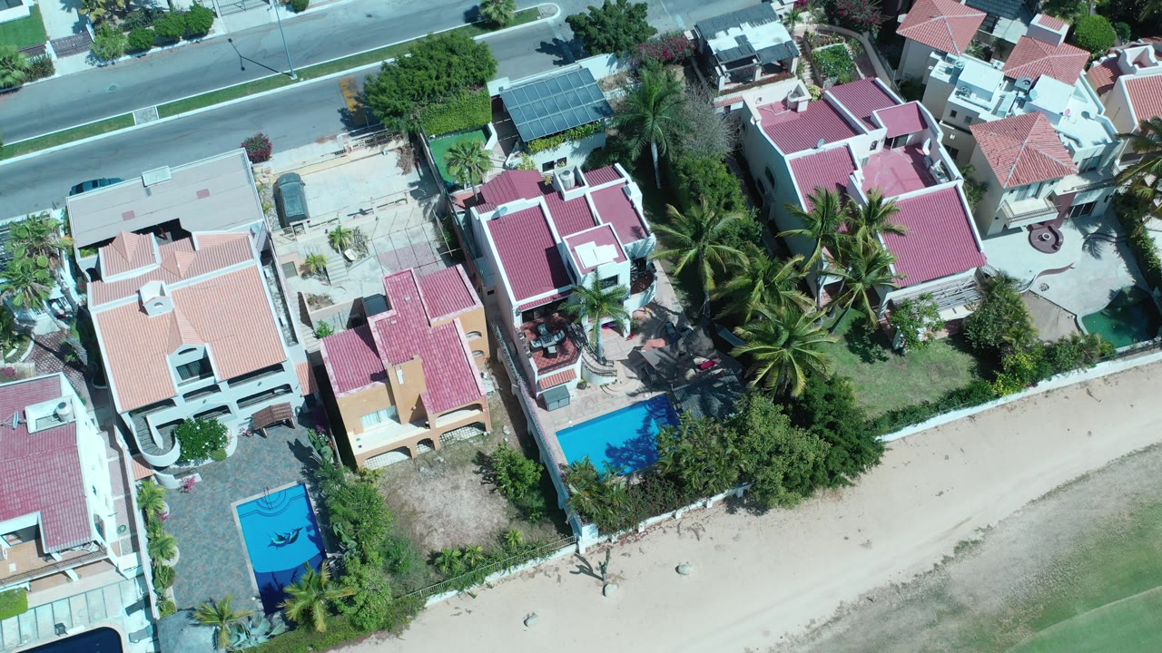 Opening overhead frame in Los Cabos, Mexico showing red and peach roofed homes, blue pools, palms, a paved street, and no visible people.