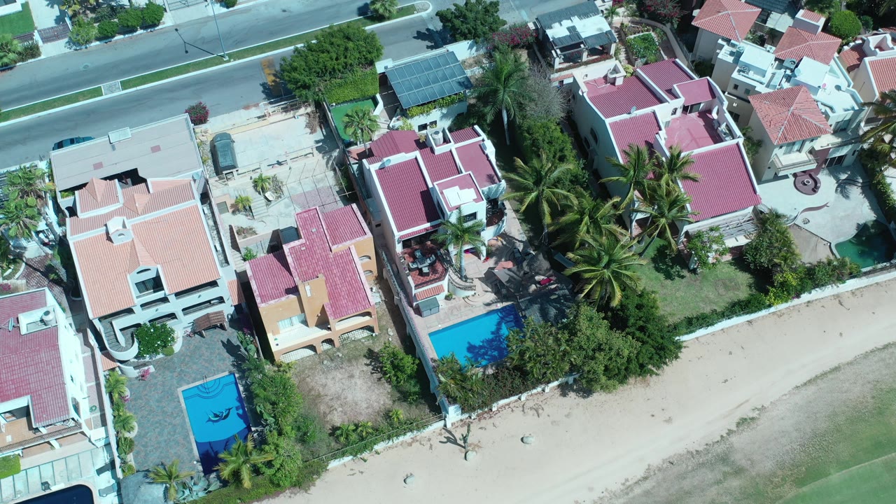 Second overhead frame in Los Cabos, Mexico with the street along the top edge, clustered red roofs, two blue pools, palms, and no visible people.