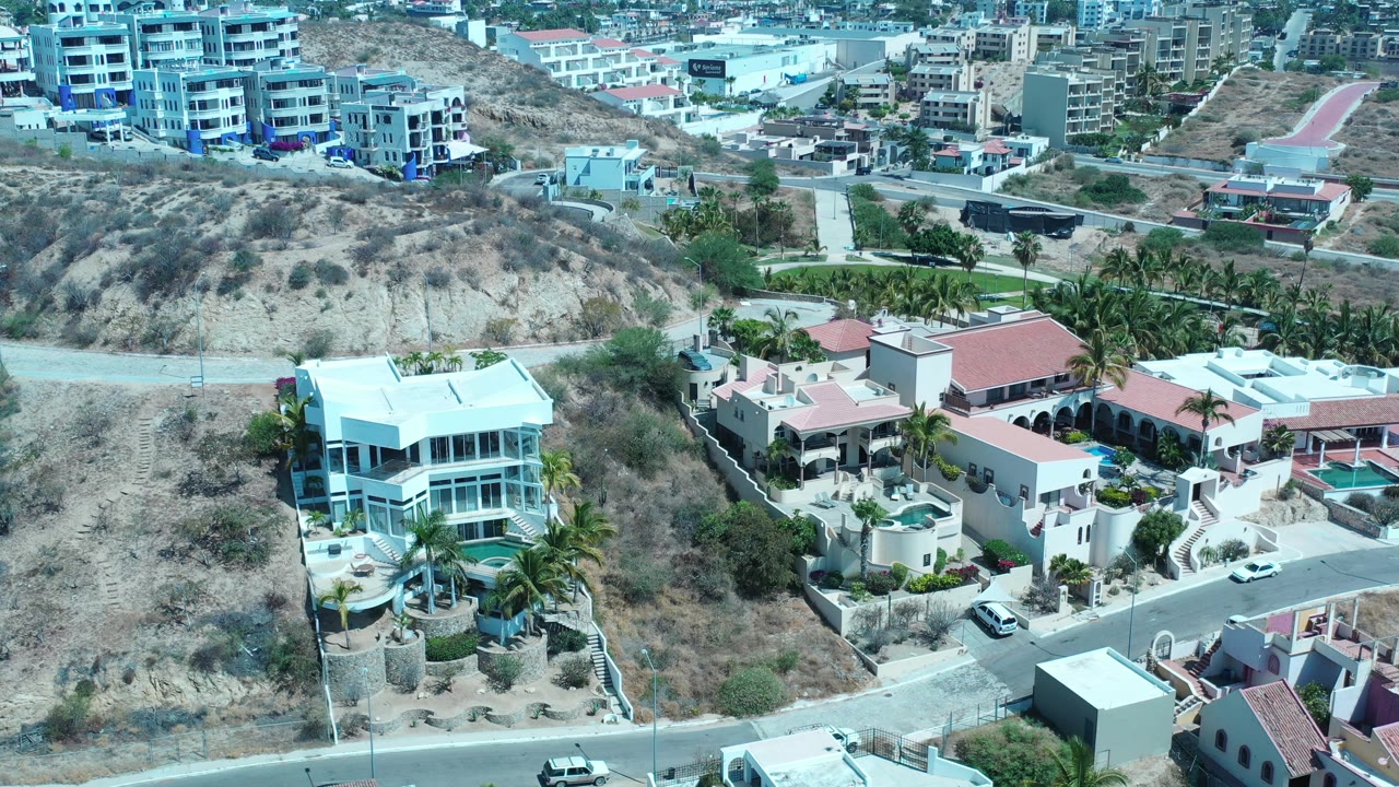 Opening aerial view over Los Cabos homes, a dry hillside road, palm trees, distant buildings, bright sky, and no visible people.