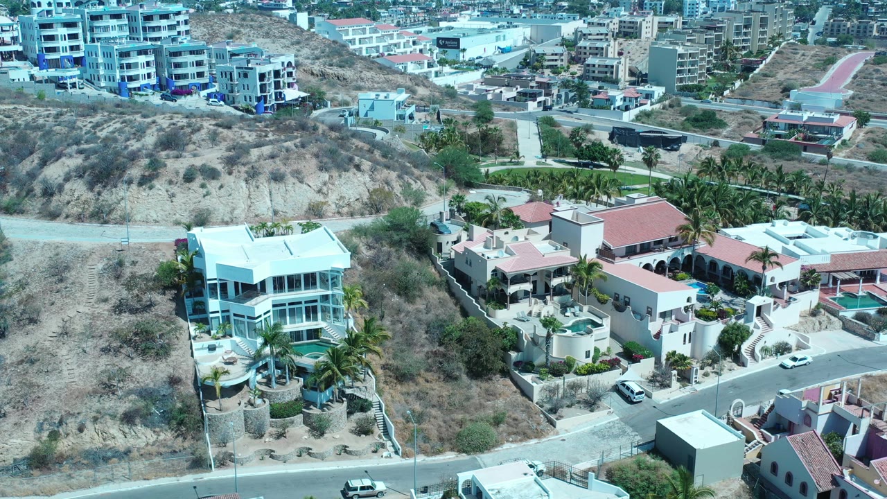 Aerial frame in Los Cabos centered on a white multi-story house beside a dry slope, with roads, palms, distant buildings, and no visible people.