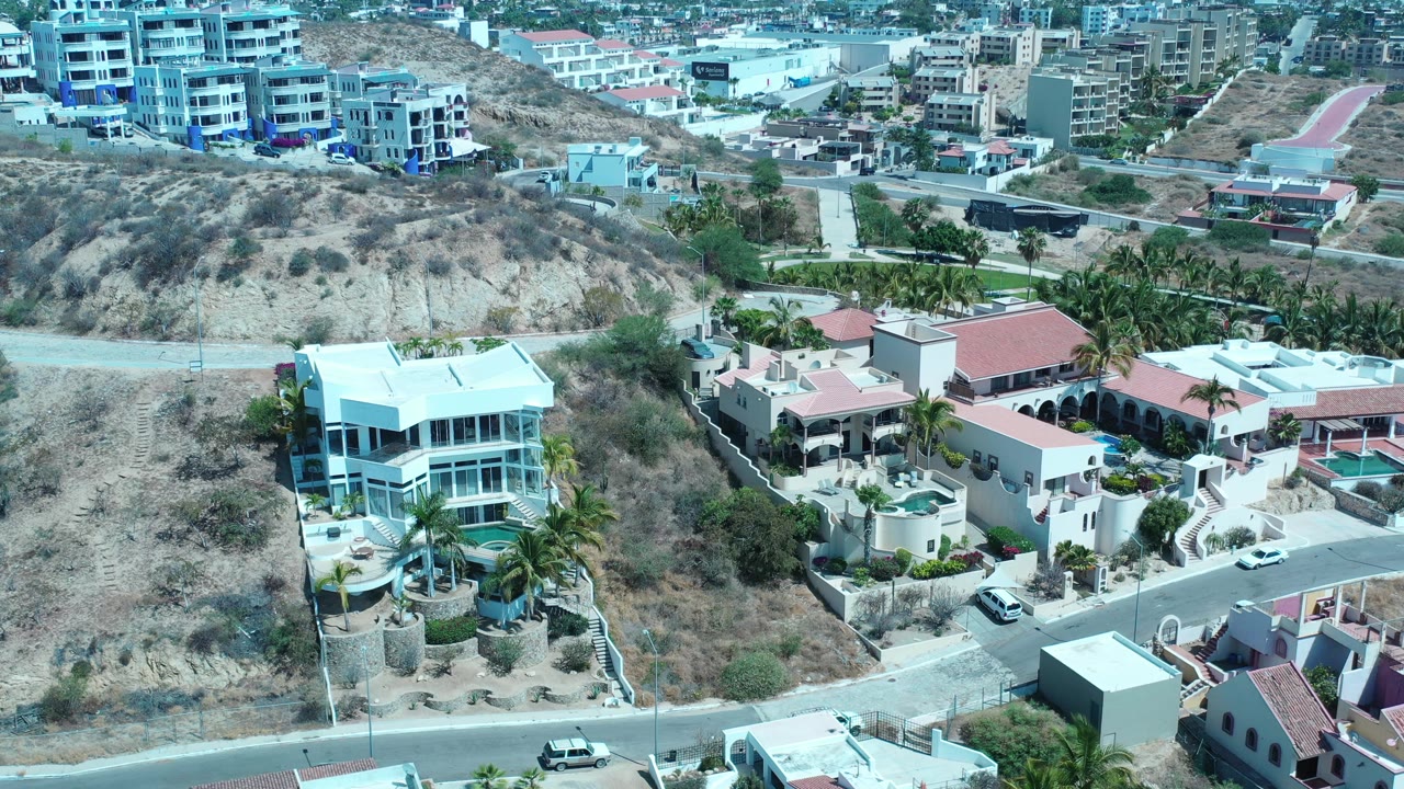 Drone view of homes and a scrub hillside near larger buildings.