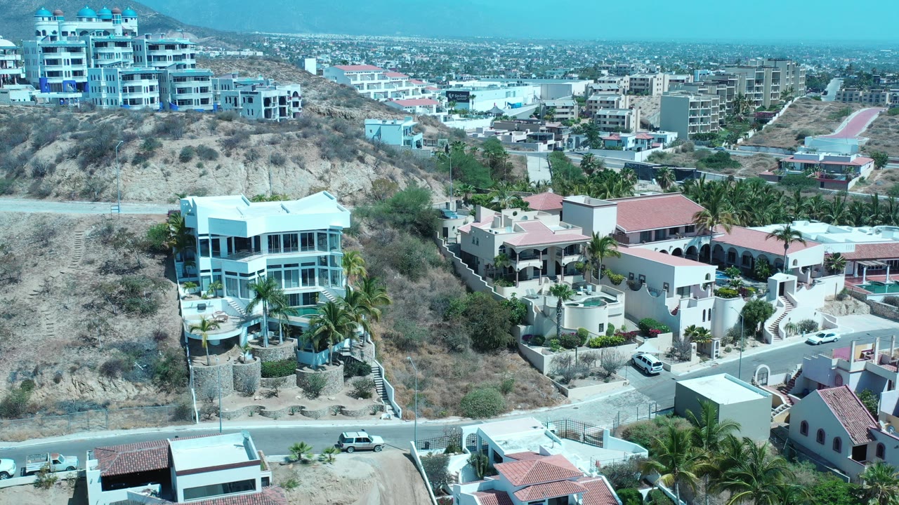 Elevated aerial frame over Los Cabos, Mexico, showing white hillside houses, dry scrub slopes, palms, roads, distant buildings, and no visible people.