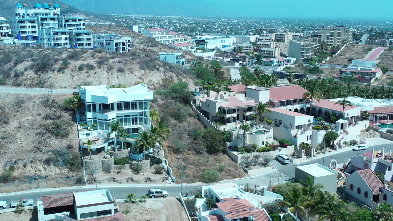 Aerial view in Los Cabos, Mexico, with a large white house near the lower left, palm trees, dry hillside lots, red roof homes, and no visible people.