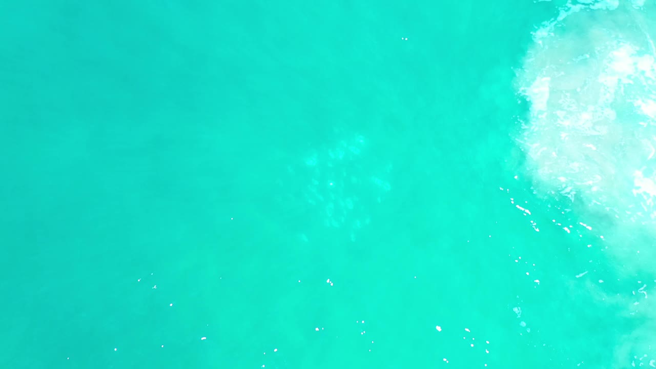 Overhead view of turquoise-green water in Los Cabos, Mexico, with faint wave shadows and a bright sun reflection near the upper right, no visible people.