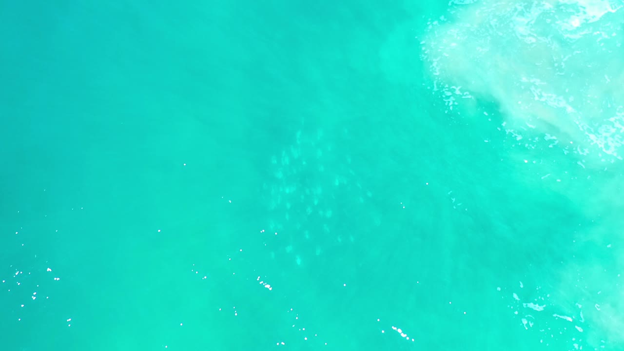 Overhead view of turquoise ocean in Los Cabos, Mexico, with a pale patch of wave foam near the upper right and no visible people.