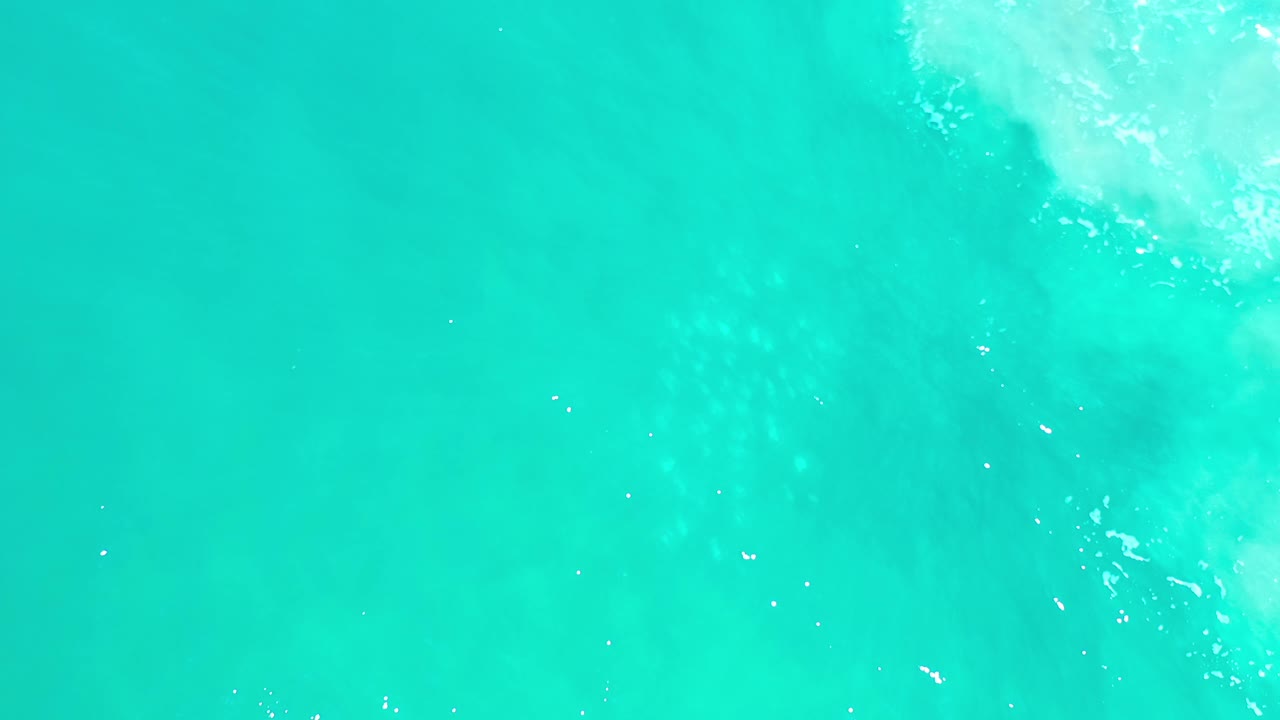 Overhead Los Cabos ocean frame showing mostly uninterrupted teal water, faint subsurface shadows, small bright flecks, and no visible people.