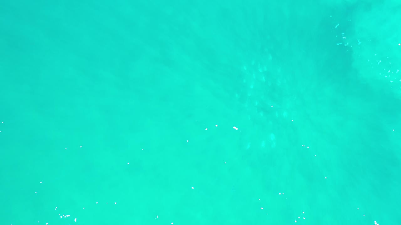Top-down view of clear turquoise-green sea in Los Cabos, Mexico, with faint darker patches below the surface and no visible people.