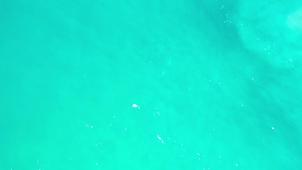 Overhead Los Cabos sea frame with bright cyan water, small white glints on the surface, and no visible people.