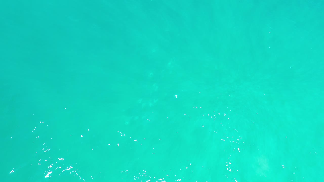 Turquoise ocean water in Los Cabos fills the frame with faint pale patches beneath the surface and small white sun glints, with no visible people.