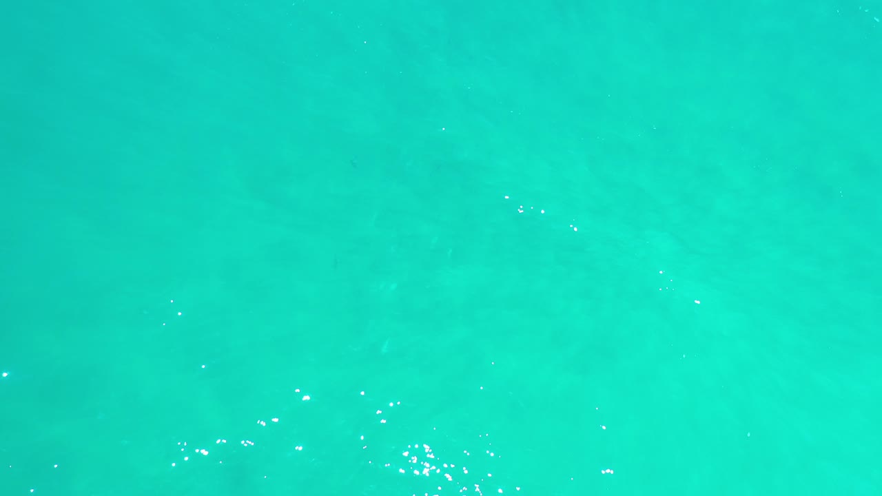 Overhead view of turquoise-green ocean water in Los Cabos, Mexico, with faint submerged patches, scattered highlights, and no visible people.