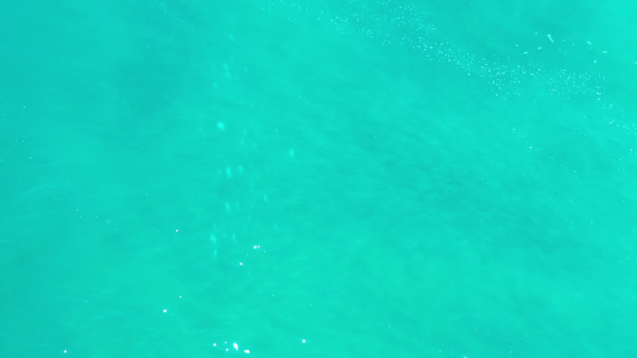 Overhead view of green-turquoise ocean water in Los Cabos, Mexico, with faint darker patches and tiny white surface flecks, no visible people.