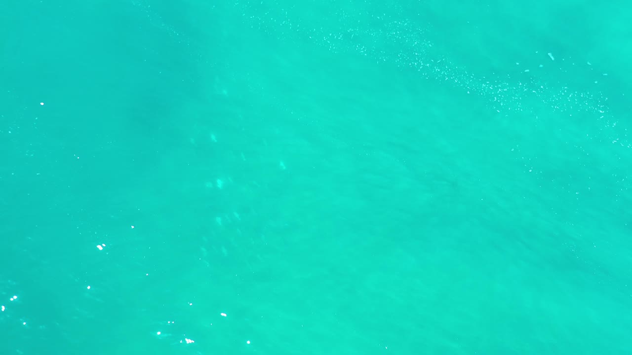 Overhead Los Cabos water frame with green-turquoise color, cloudy darker areas beneath the surface, sparse foam flecks, and no visible people.