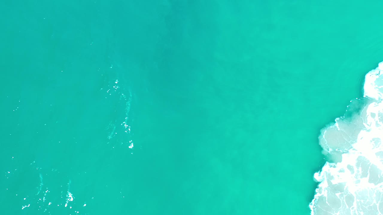Overhead view of turquoise-green sea in Los Cabos, Mexico, with white surf pushing in from the right edge and no visible people.