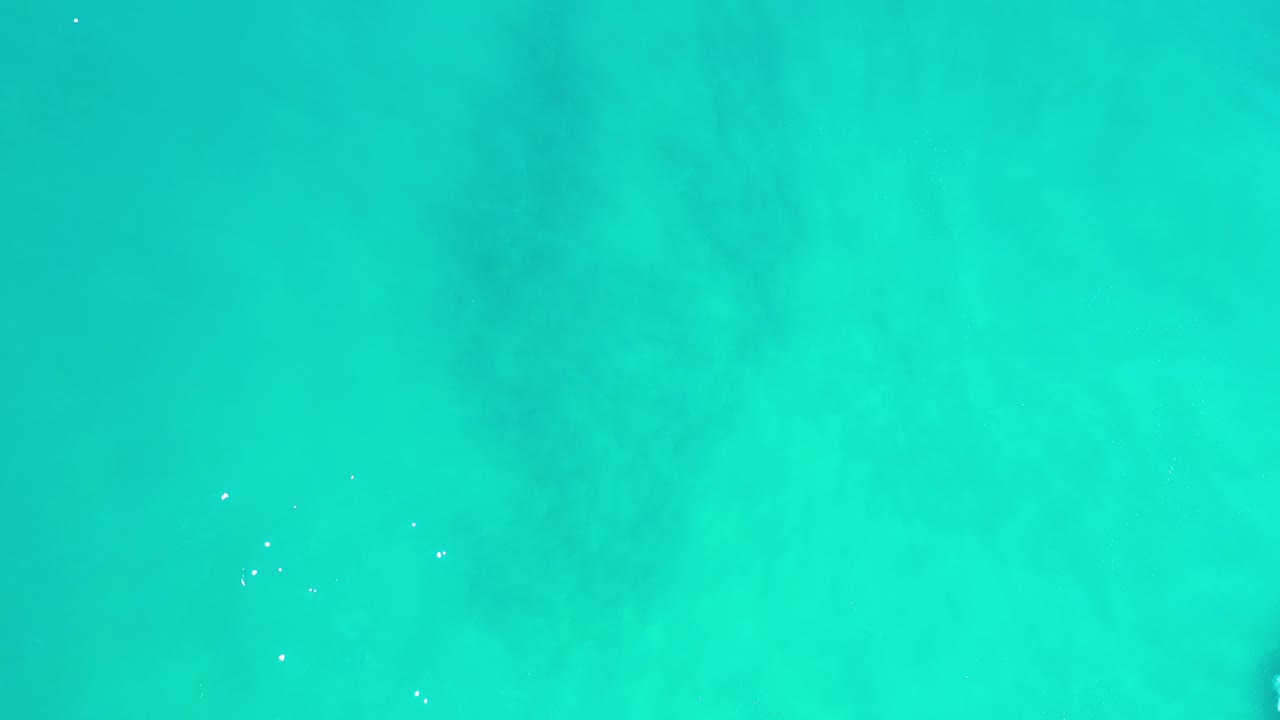 Overhead Los Cabos ocean frame with blue-green water, faint darker bands below the surface, tiny white foam spots, and no visible people.