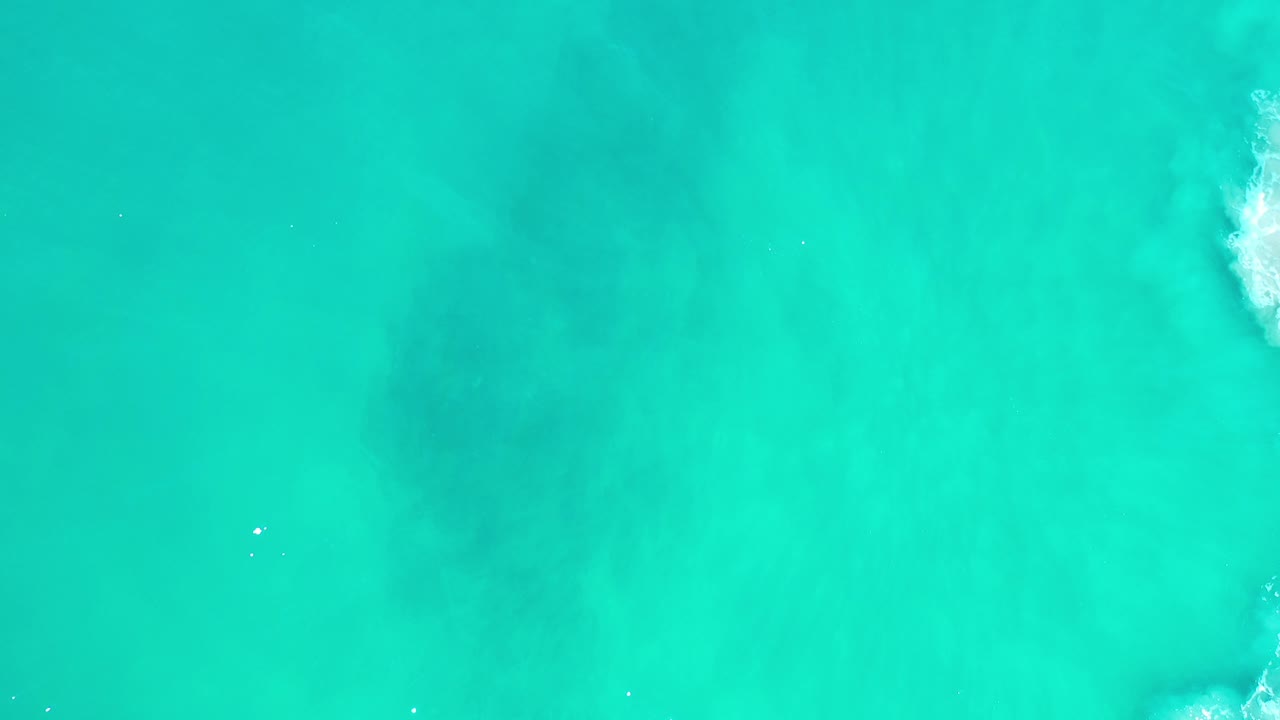 Overhead view of green-turquoise ocean water in Los Cabos, Mexico, with a soft dark patch below the surface, a small white wave break near the upper right, and no visible people.