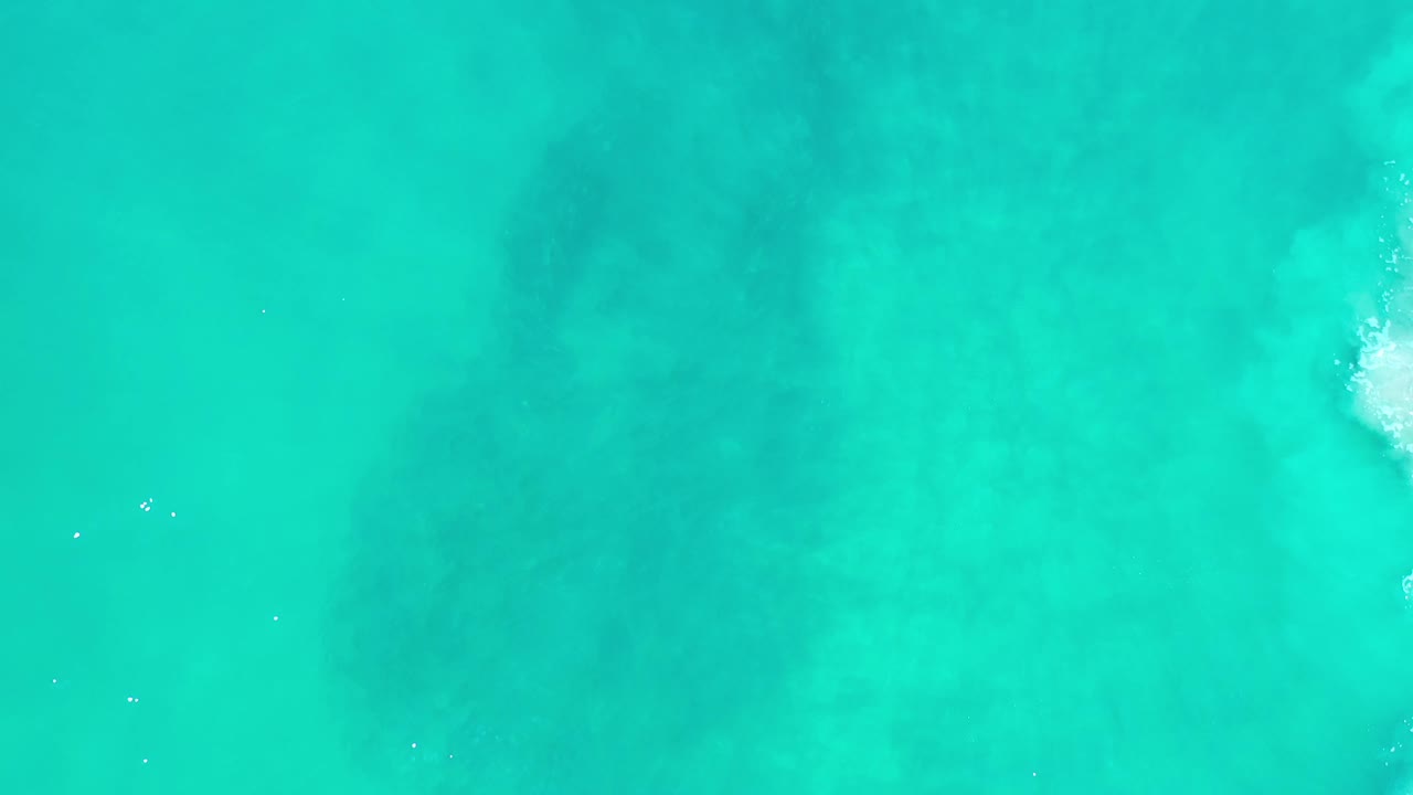 Aerial view of blue-green Los Cabos ocean water with a small white foam patch entering near the right edge and no visible people.