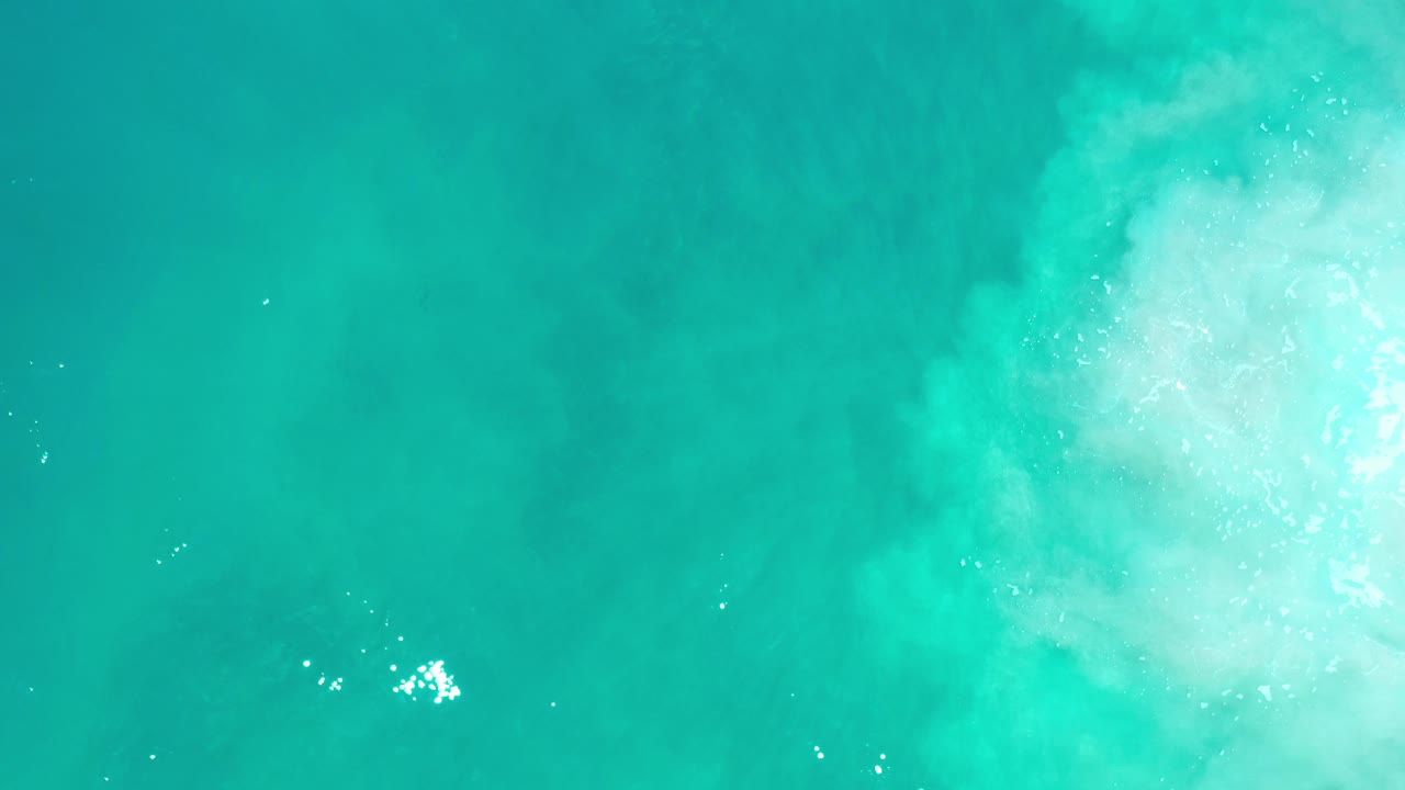 Overhead view of cyan ocean water in Los Cabos, Mexico, with pale foam gathered near the upper right and no visible people.
