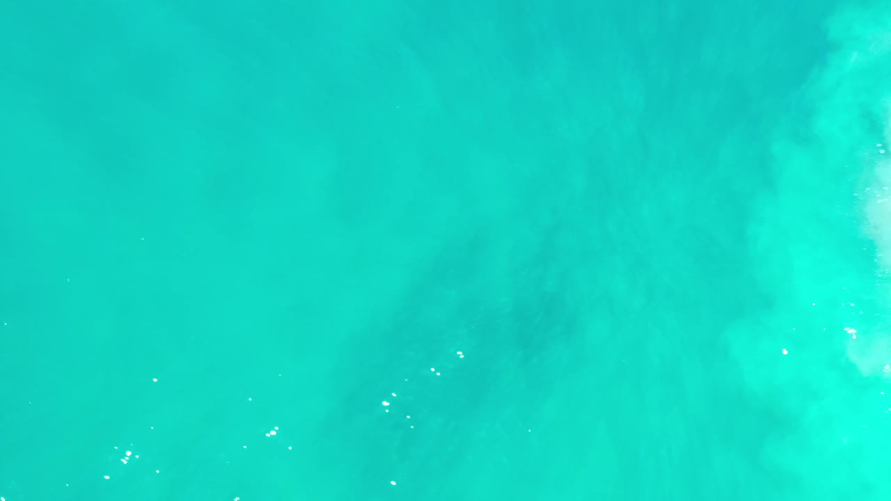Overhead Los Cabos ocean frame with blue-green water, diffuse pale wave clouding through the center, tiny white surface glints, and no visible people.