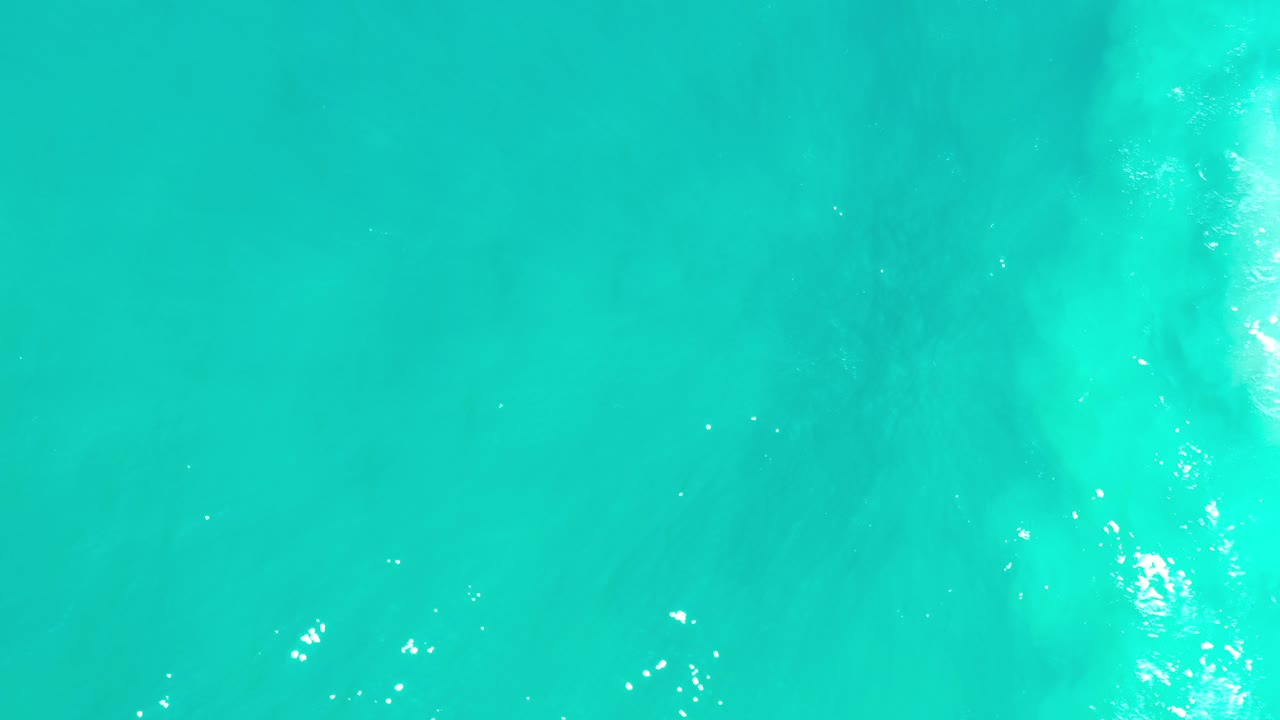 Overhead view of turquoise water in Los Cabos, Mexico, with faint pale underwater patches, scattered sun glints, and no visible people.