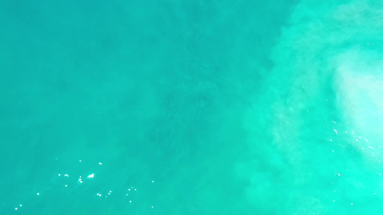 Overhead Los Cabos ocean frame with soft white foam along the upper edge, scattered sun glints, and no visible people.