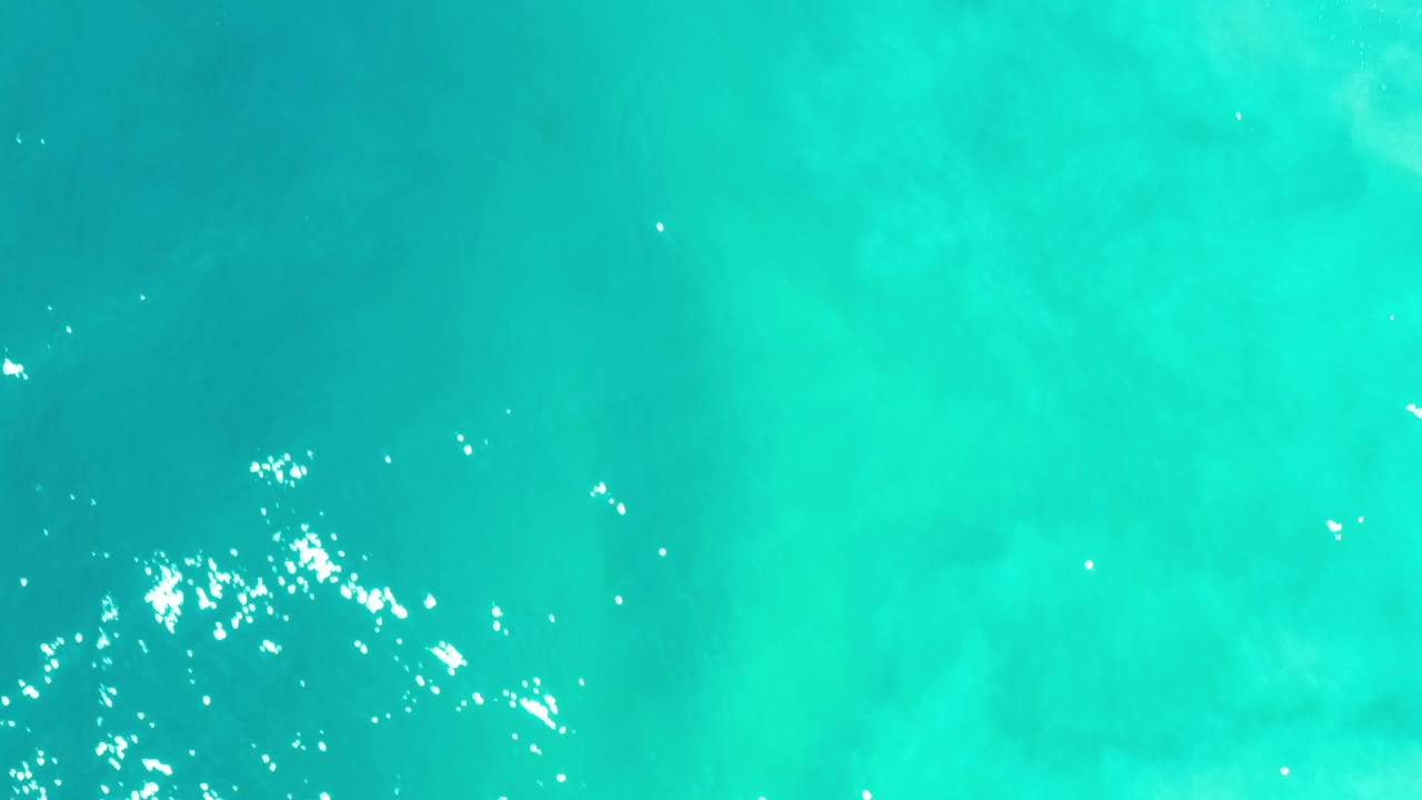 Top-down view of turquoise sea in Los Cabos, Mexico, with scattered white sun glints near the upper left and no visible people.