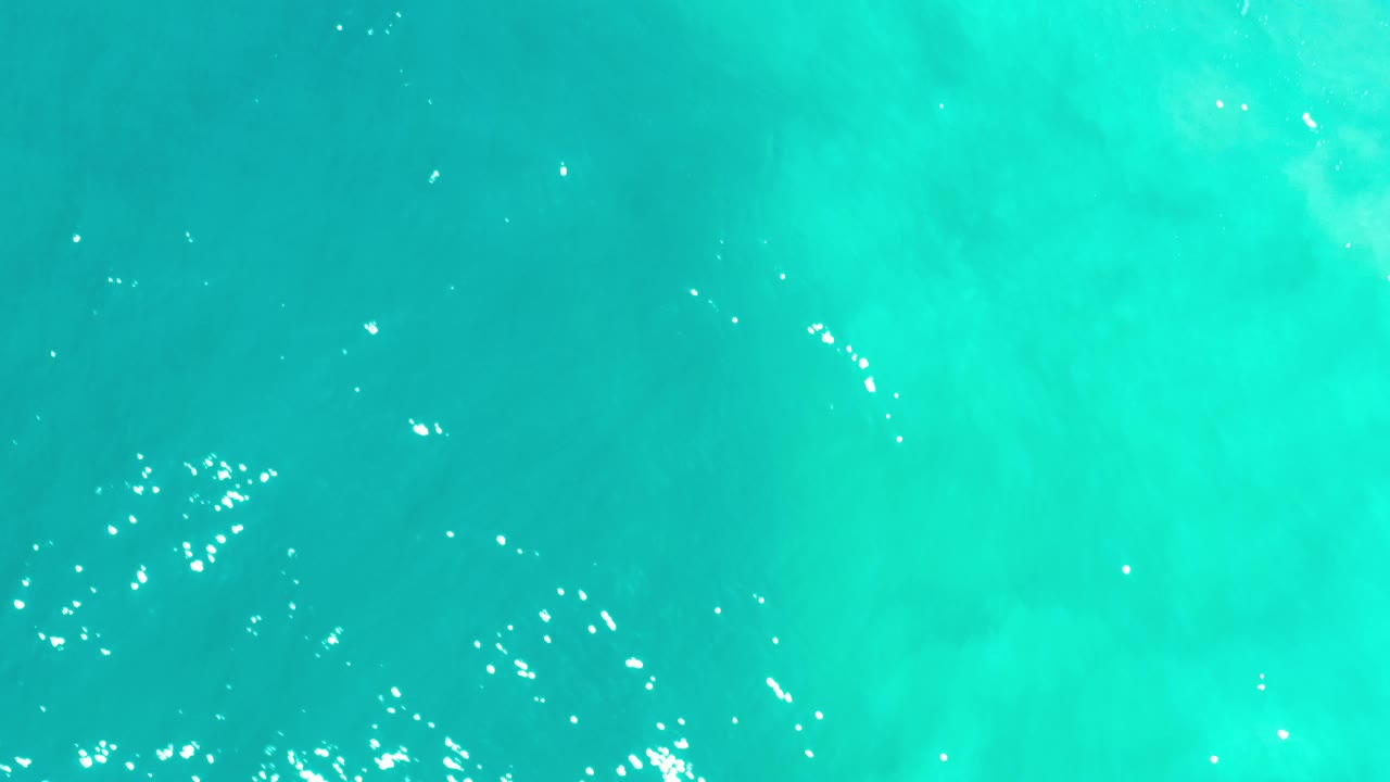 Top-down turquoise water in Los Cabos, Mexico, with darker depth patches, scattered surface sparkles, and no visible people.
