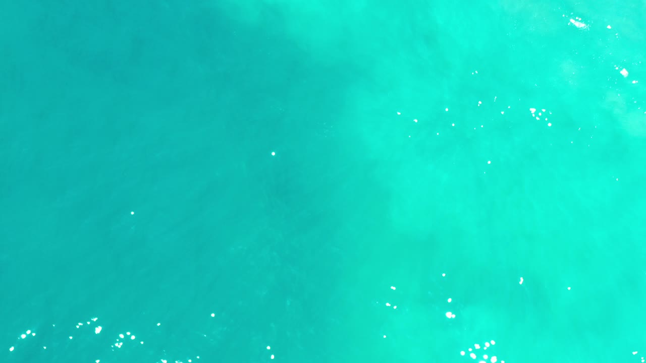Top-down view of turquoise ocean water in Los Cabos, Mexico, with scattered white sun reflections near the lower left and no visible people.