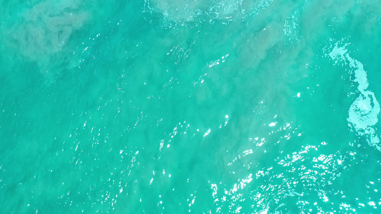 Overhead view of turquoise-green sea in Los Cabos, Mexico, with sunlit ripples and a small white foam patch near the right side, no visible people.