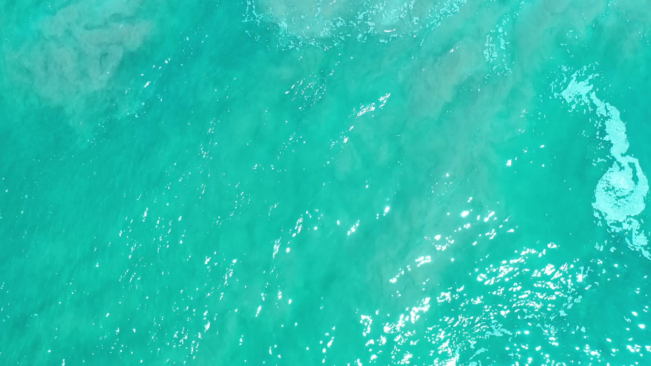 Aerial view of green sea in Los Cabos, Mexico, with brighter sun glitter across the lower water and a narrow white foam streak near the upper right, no visible people.
