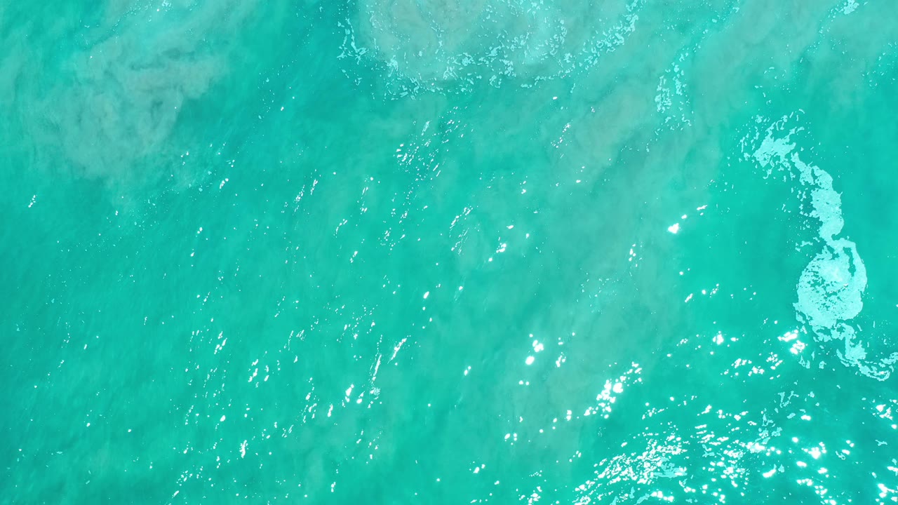 Turquoise water seen from above with ripples and a white foam patch on the right.