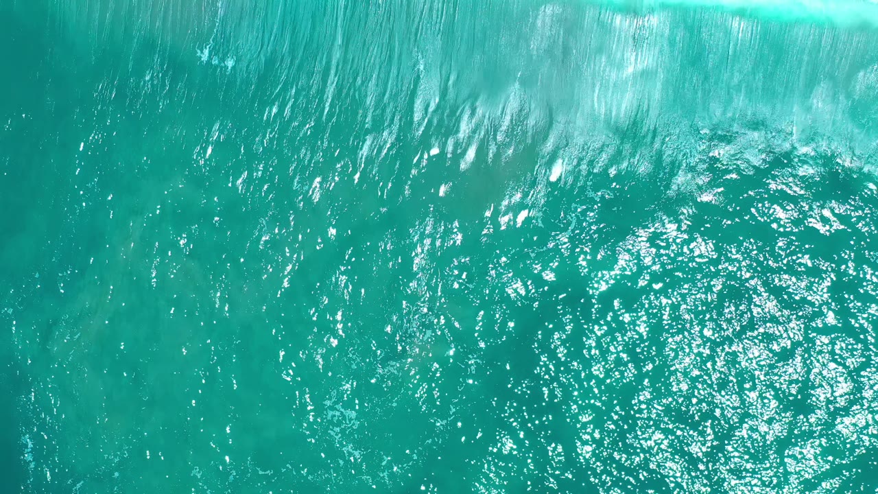 Aerial frame over blue-green water in Los Cabos, Mexico, with concentrated sun glitter across the rippled surface and no visible people.