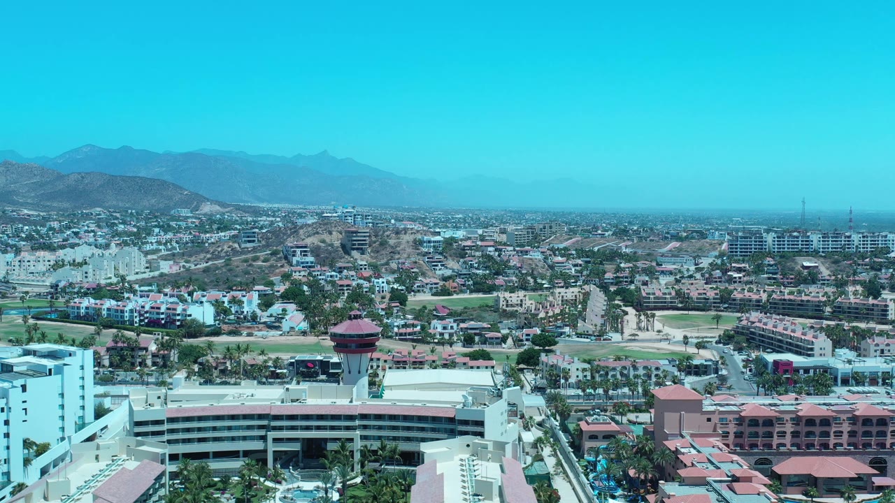Early aerial view in Los Cabos, Mexico, with the resort tower slightly lower in frame, roads and palm rows crossing the foreground, dense buildings beyond, mountains, and no visible people.