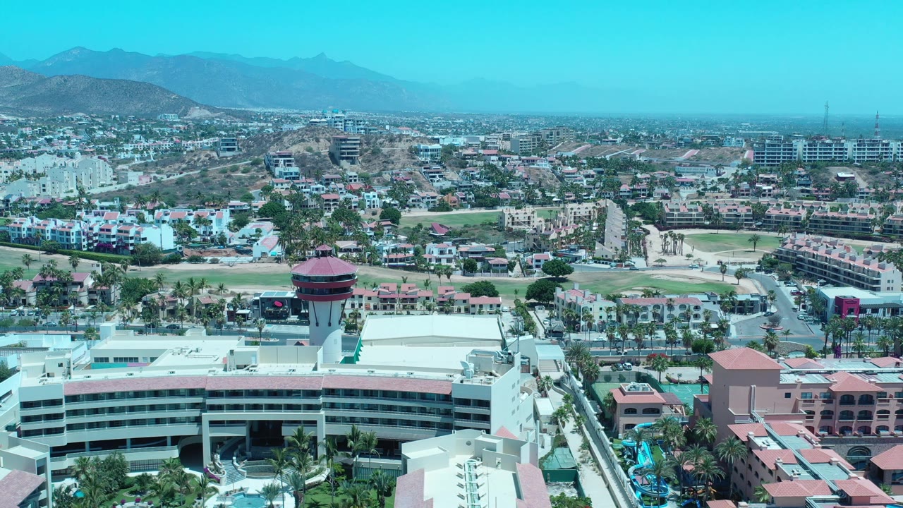 One second in, the Los Cabos aerial holds the red-roofed tower near the lower center, with roads, fairways, modern low-rise buildings, dry hills, and no visible people.