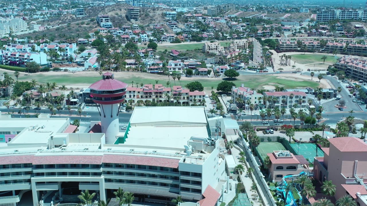 Opening aerial frame over Los Cabos, Mexico, showing a round red-roofed tower beside white buildings, roads, palm trees, green sports courts, and no visible people.
